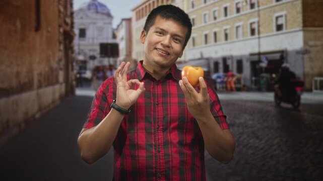 Man holding persimmon in right hand and making ok sign with left hand on a cobblestone street; simple joy.