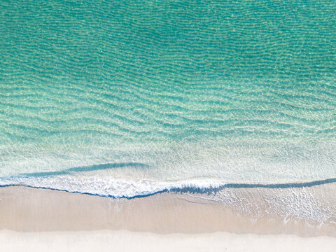 Aerial view of gentle waves on sandy beach with clear turquoise water.