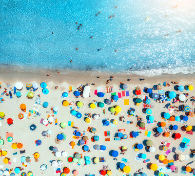 Aerial view of colorful umbrellas on sandy beach, swimming people in blue sea at sunset in summer in Sardinia, Italy. Tropical landscape with turquoise transparent water. Travel and vacation. Top view