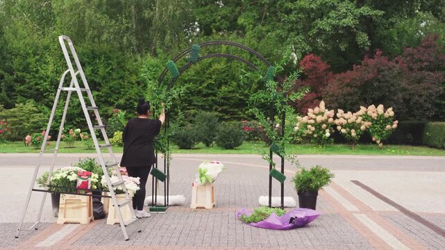 Wedding floral setup. Elegant garden backdrop with floral arrangements and lush greenery. Calm summer outdoor setting with florist creating wedding decorations and floral installations