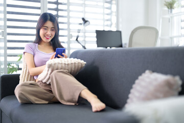 Obraz premium Asian adult woman relaxing comfortably on sofa using smartphone browsing social media happy casual home living room modern lifestyle communication