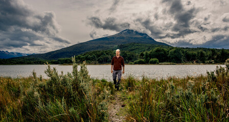 Fototapeta premium Hiker walking along a trail toward a dramatic mountain peak and lake view under overcast sky, near Río Lapataia, Tierra del Fuego.