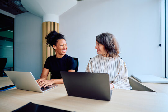 Coworkers smiling and communicating during a meeting, working on laptops in a contemporary office environment