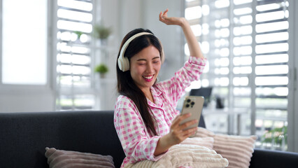 Fototapeta premium Asian adult woman listening music headphones using smartphone enjoying relaxing time cozy home modern living room leisure lifestyle happiness comfort