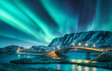 Naklejka premium Bridge and northern lights over snowy mountains. Lofoten islands, Norway. Aurora borealis and reflection in water. Winter landscape with starry sky, polar lights, rocks, road, sea, city illumination