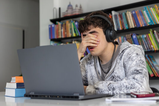 Stressed and exhausted teenage student rubbing tired eyes while studying with laptop and books, preparation for difficult exams, academic pressure, ADHD concept.