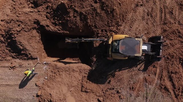 Aerial Top Down View of an Excavator Digging a Foundation Pit with a Construction Worker Directing