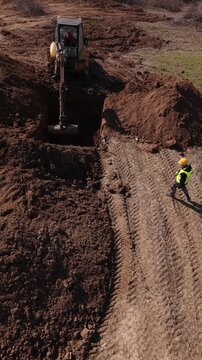 Vertical drone footage of a yellow excavator digging a deep rectangular trench in the soil. A construction worker wearing a hard hat and safety vest stands on the edge of the pit.