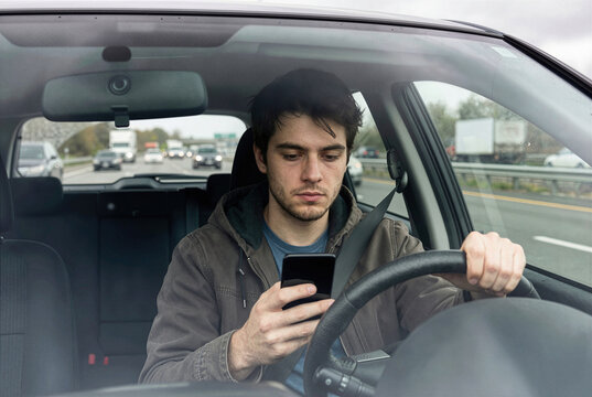 Young man distracted driving while using smartphone, male driver looking at mobile phone behind the steering wheel, dangerous behavior inside car on highway, road safety concept.