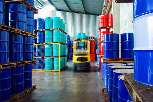 Industrial warehouse storage with rows of stacked blue, teal, and red oil barrels and a blurred yellow forklift moving through the aisle. Chemical logistics and fuel distribution concept.