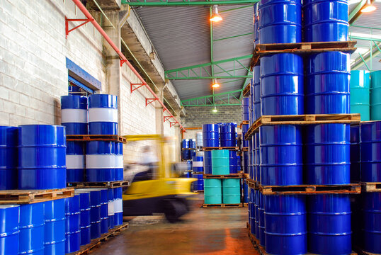 Industrial warehouse storage with rows of stacked blue, teal, and red oil barrels and a blurred yellow forklift moving through the aisle. Chemical logistics and fuel distribution concept.