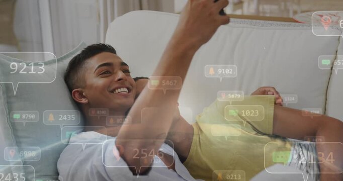 Raising smartphone, blue-shirted man and partner in yellow tee framing selfie on sofa smiling at ale