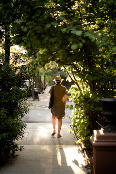 A trendy woman in a dress and hat walks down a street at sunset.