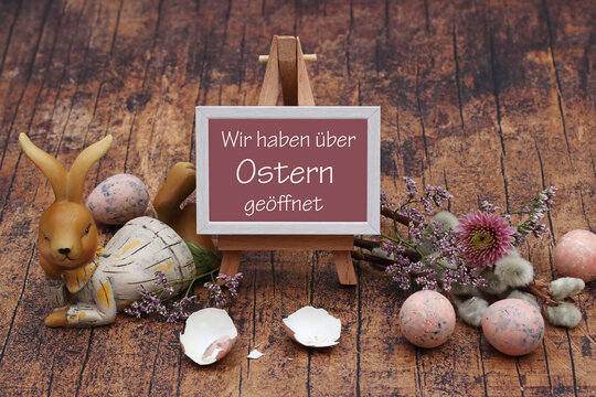 Liegende Hasenfigur neben kleiner Staffelei-Schild mit dem Text 'Wir haben &uuml;ber Ostern ge&ouml;ffnet', umgeben von zerbrochenen Eierschalen, Osterzweigen und dekorativen Eiern auf Holzboden.