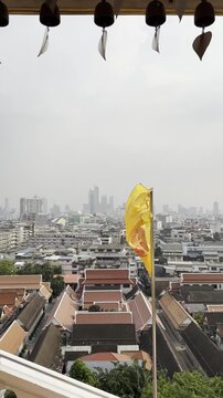 Yellow Buddhist flag with golden dharma wheel waving over temple rooftops with panoramic Bangkok city skyline and hanging bells in spiritual morning atmosphere