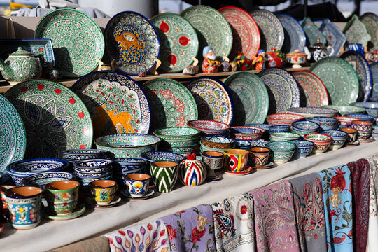 Colorful ceramic plates bowls, cups displayed on market stall Siab bazaar as oriental souvenirs. Sunny day, vibrant handmade pottery - floral, geometric patterns, scarfs. Samarkand, Uzbekistan.