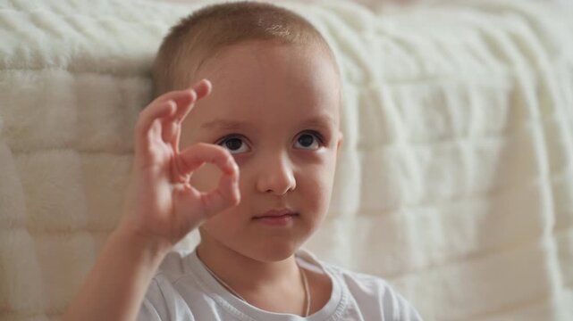 Mischievous boy making okay sign in bedroom. Young child with toy displaying playful confidence in cozy room. Cheerful young boy shows affirming gesture with toy in comfortable bedroom setting