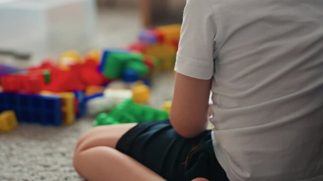 White boy sitting among colorful blocks on carpet, pensive profile beside striped blanket, quiet indoor bedroom scene, closeup of back and head, contemplative pause during playtime, childfocused mood