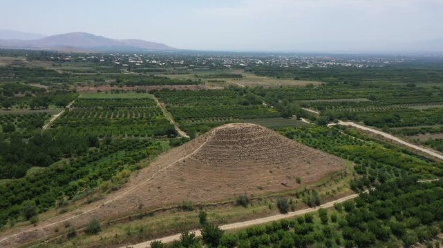 Aerial drone footage of a mysterious ancient stepped earthen pyramid, possibly a burial mound or kurgan, beautifully situated amidst lush agricultural fields and orchards in a scenic valley.

