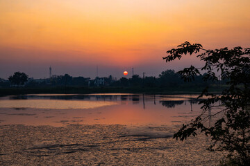 Fototapeta premium Dusty and cloudy sunrise silhouetting the skyline of Tankal reflected in the still water of Tankal Lake, Gujarat, India