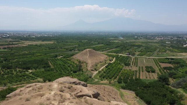 Aerial drone footage of a mysterious ancient stepped earthen pyramid, possibly a burial mound or kurgan, beautifully situated amidst lush agricultural fields and orchards in a scenic valley.

