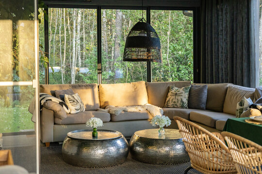 Cozy modern sunroom with large beige corner sofa, patterned cushions, soft throw, woven pendant lamp, metallic hammered coffee tables with white flowers, floor-to-ceiling windows with forest view.