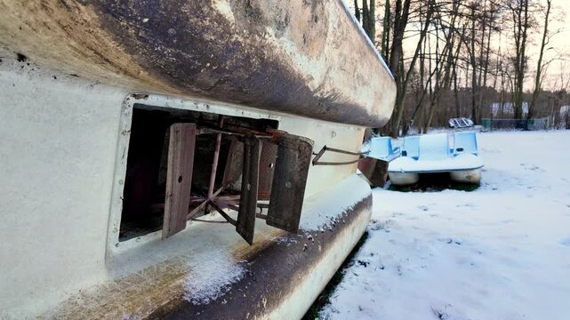Old paddle wheel mechanism of a pedal boat on the lake shore in winter. Damaged boat hull with metal paddles against a snowy landscape, forest, and blue pedal boat background.