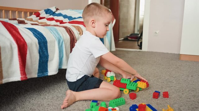 Boy building vibrant toy train. Energetic youngster putting together multicolored train pieces on carpet. Playful boy creating vivid toy train while relaxing barefoot beside striped bed
