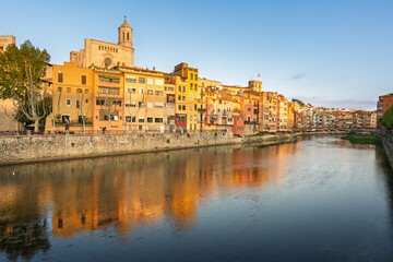 Fototapeta premium Girona cityscape featuring the colorful historic houses lining the Onyar River, with the ancient Cathedral of Girona rising above the rooftops under a clear sky, reflecting in the water