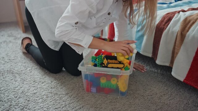 Mom kneeling by bed gathering toys from plastic bin, reaching to pick up colorful blocks and place container beside blanket, soft carpet and bedroom textiles create warm home cleanup scene, careful
