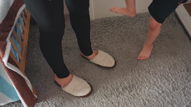 Mom and son morning dressing routine moment, tender bedroom scene with mother helping barefoot toddler into slippers on carpet beside crib, gentle bending, guiding hands, warm cozy lighting, domestic