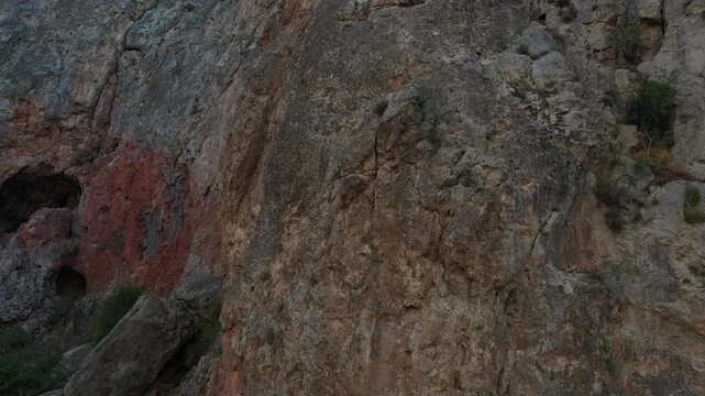 Slowly zooming into a mysterious, dark cave entrance set within a massive, rugged, reddish-brown rocky cliff face, highlighting the harsh, ancient geological formation in a remote natural landscape.

