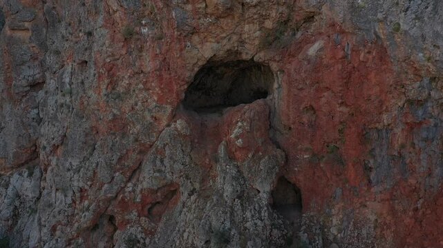 Slowly zooming into a mysterious, dark cave entrance set within a massive, rugged, reddish-brown rocky cliff face, highlighting the harsh, ancient geological formation in a remote natural landscape.

