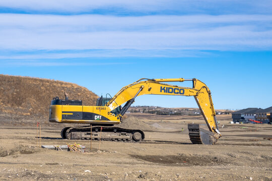 Calgary, Alberta, Canada. March 27, 2026. A large yellow CAT excavator branded with KIDCO logos sits at a suburban residential land development site.