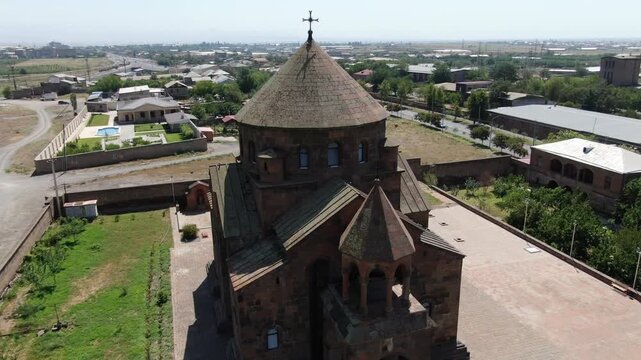 Aerial drone footage circling a magnificent ancient Armenian church made of stone with a distinctive conical dome, situated in a town surrounded by trees on a sunny day.

