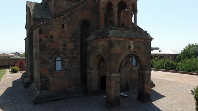 Aerial drone footage circling a magnificent ancient Armenian church made of stone with a distinctive conical dome, situated in a town surrounded by trees on a sunny day.

