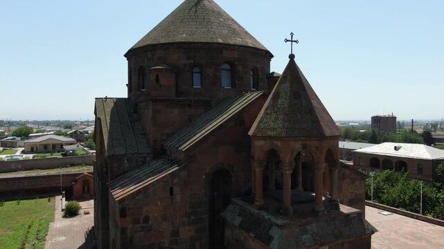 Aerial drone footage circling a magnificent ancient Armenian church made of stone with a distinctive conical dome, situated in a town surrounded by trees on a sunny day.

