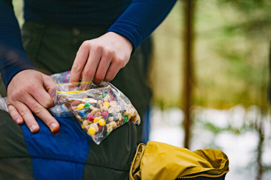 A young man opens a plastic bag of raisins, peanuts and chocolate candies while backpacking in winter