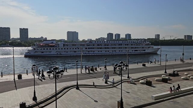 MOSCOW - JUL 26 , 2025: Large cruise ship is docked at a pier. The pier is lined with street lamps and benches. People are walking around and sitting on the benches.