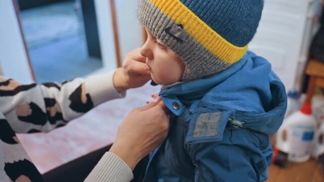 Mom fastening sons winter jacket at doorway, closeup hands adjusting zipper and knit hat, soft morning light through window, attentive parent preparing toddler for outing, cozy blue coat and gentle
