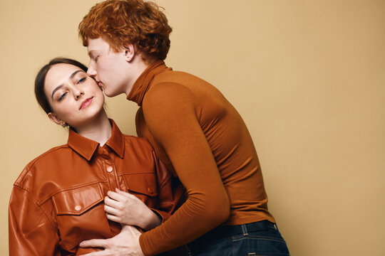 Couple with man kissing woman on cheek for love and affection. Young couple in tender moment wearing brown clothes on beige background with closed eyes and intimacy.