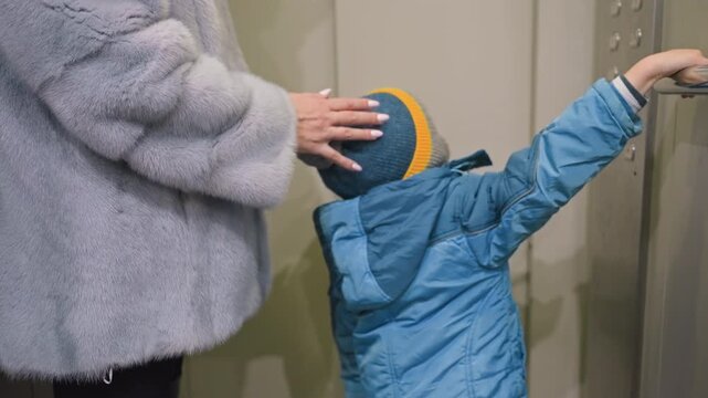 Mom and son at elevator, mother adjusts sons knit hat while child reaches to press button, holding small toy, soft gray coat and blue puffer jacket, gentle touch and guiding hand, intimate family