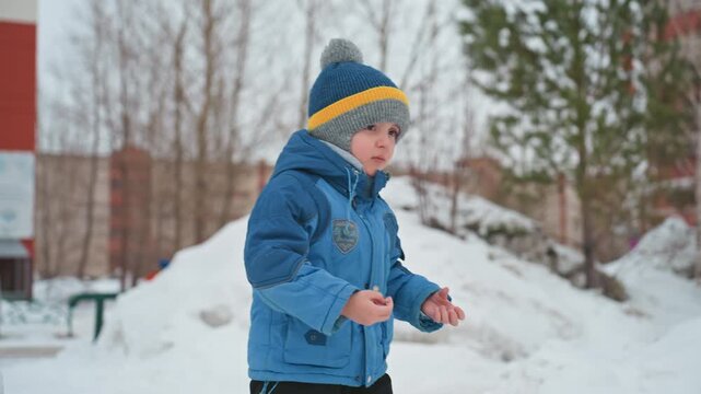 Son tossing snowball near snowy mound, white boy in blue jacket and knit hat lifts snow into air, energetic motion, footprints across park, urban playground backdrop, curious independent play, joyful