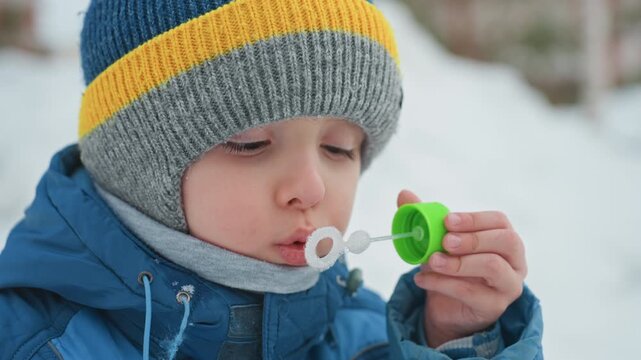 Child in winter blowing delicate snow bubbles. Closeup of child in blue coat creating shimmering snow bubbles. Young child bundled in winter gear blowing iridescent bubbles in cold snow scene