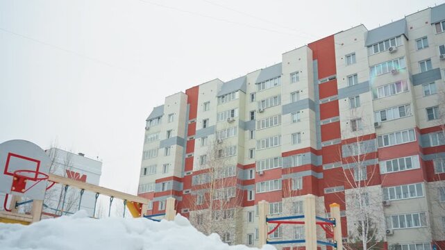 Son blowing bubbles in snowy courtyard near apartment buildings, bundled in blue coat and knit hat, breath visible in cold air, bubbles reflecting overcast sky, playground hoop and snowbanks framing