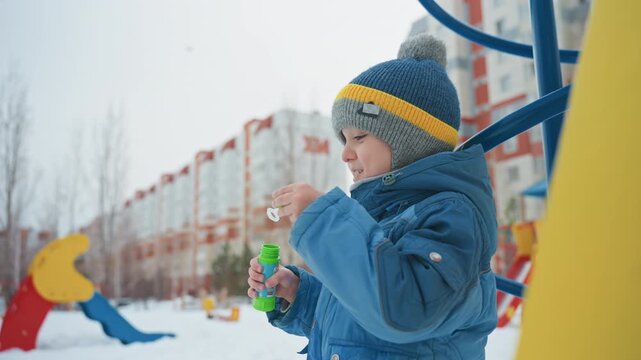 Smiling white son blowing large bubble near colorful playground slide, bundled in blue coat and knit hat, breath visible in cold air, wand creating shimmering sphere that floats past urban buildings,