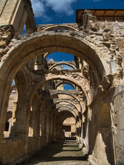 Fototapeta premium Long perspective view of ancient stone archways and cloisters - Architectural ruins of a historic monastery or abbey with sunlight streaming through the open arches and blue sky above.