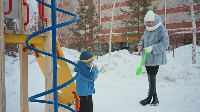 White mom and son playing with green shovel in snowy playground, mom in gray coat and white beanie hands bright shovel to son in blue jacket, standing near spiral climbing frame, playful winter