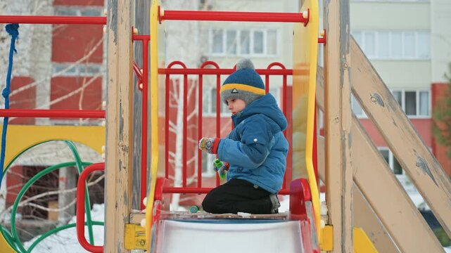 Winter playground white mom and son, bundled in blue puffer and knit beanie, little boy sits on slide inspecting snow, mother approaches from left offering gentle help, urban apartment backdrop, quiet