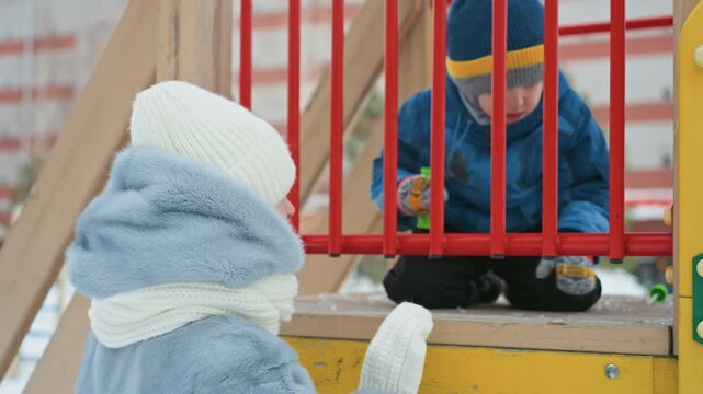 Winter playground mom with son bundled in coats and knit hats, mother watches son sitting behind red play structure bars while child inspects small toy, snowy ground and cold breath visible, tender